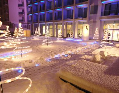 Cortile innevato con alberi di luci davanti a un edificio.