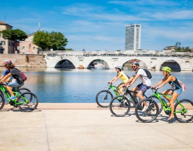 Quattro amici sorridenti pedalano lungo un fiume sotto un cielo azzurro.