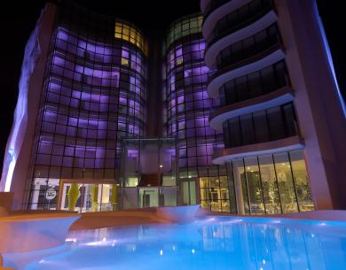 Modernes Gebäude in lila beleuchtet, spiegelt sich in einem nächtlichen Pool.