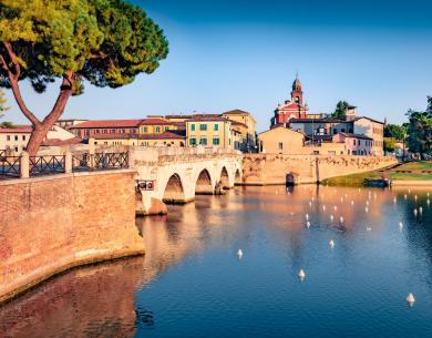 Historische Brücke über einen Fluss, umgeben von bunten Gebäuden und Bäumen.