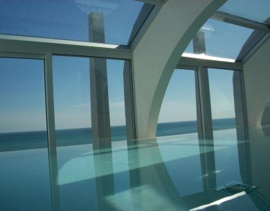 Piscina interna con vista mare, ampie finestre e arcate bianche.