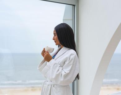 Frau im weißen Bademantel trinkt Kaffee mit Meerblick.