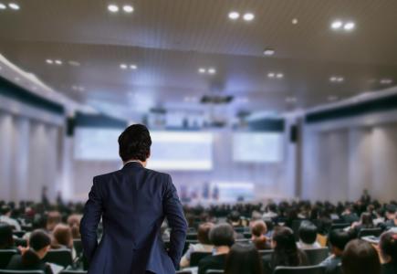 Uomo in giacca osserva platea in auditorium, atmosfera di attesa e professionalità.