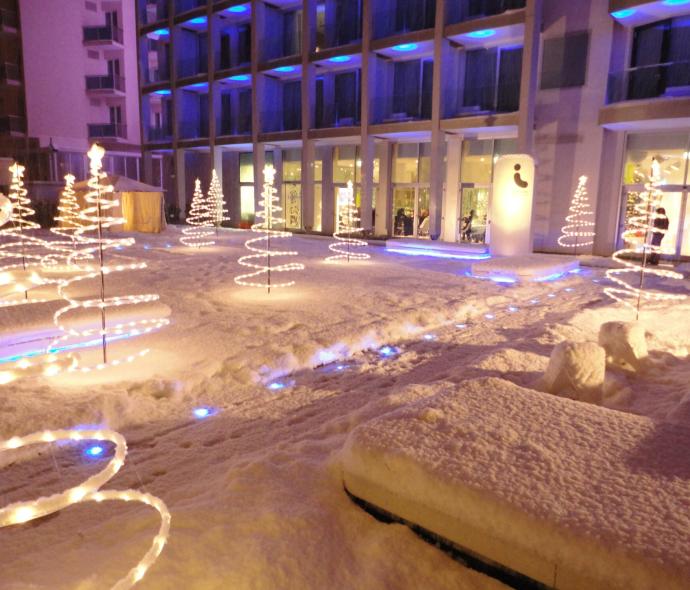 Cortile innevato con alberi di luci davanti a un edificio.