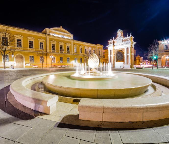 Fontana illuminata di notte in una piazza con edifici storici.
