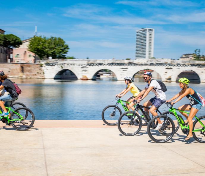 Quattro amici sorridenti pedalano lungo un fiume sotto un cielo azzurro.