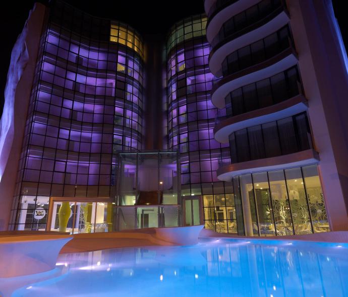 Modernes Gebäude in lila beleuchtet, spiegelt sich in einem nächtlichen Pool.