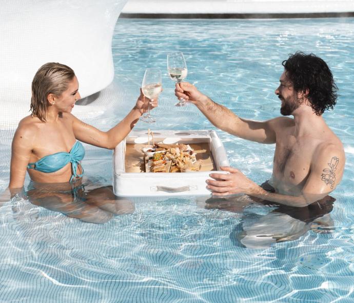 Coppia brinda in piscina con vino e snack galleggianti.