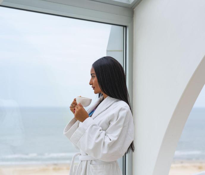 Frau im weißen Bademantel trinkt Kaffee mit Meerblick.