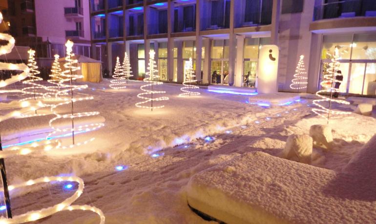Cortile innevato con alberi di luci davanti a un edificio.