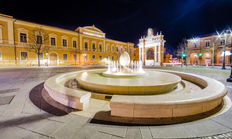 Fontana illuminata di notte in una piazza con edifici storici.