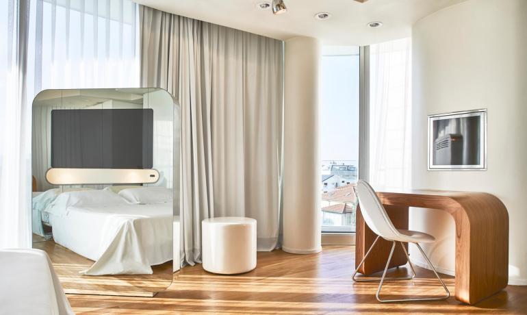 Modernes Zimmer mit Bett, Schreibtisch und großen Fenstern.