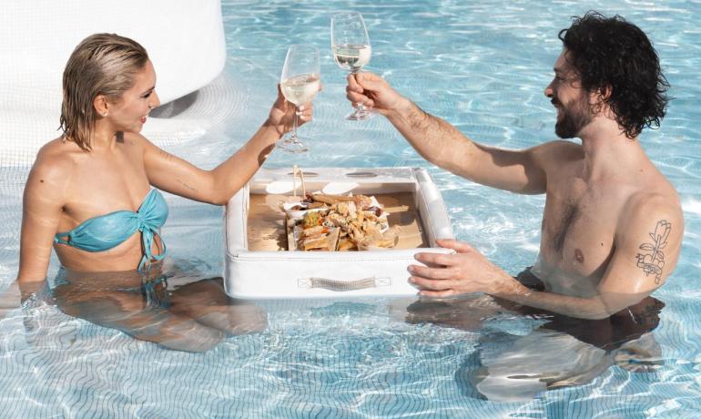 Coppia brinda in piscina con vino e snack galleggianti.