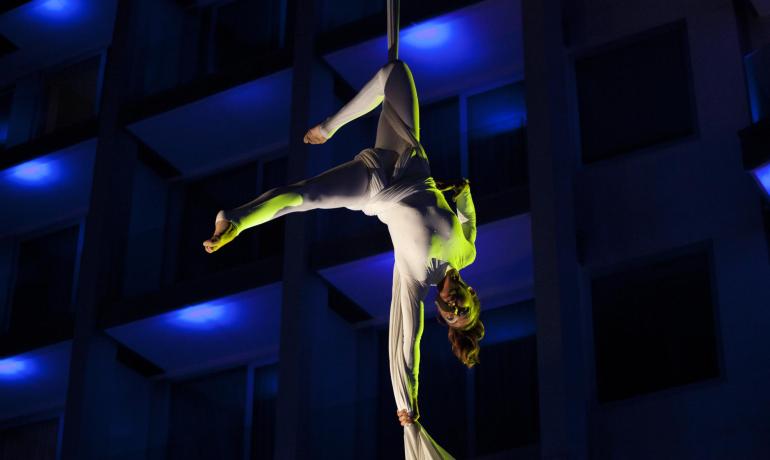 Aerial performer suspended in air, wrapped in fabric, with intense blue and green lights.