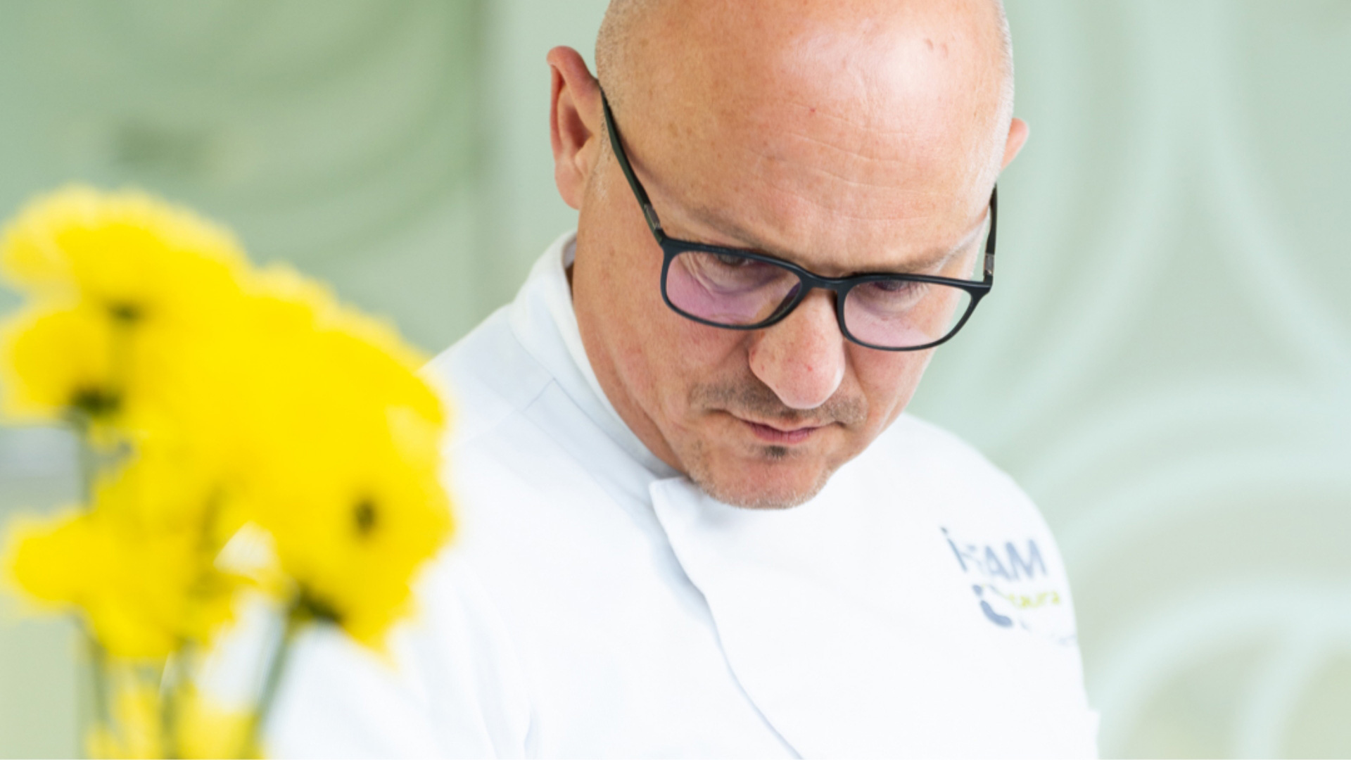 Chef concentrato con occhiali, indossa una giacca bianca, fiori gialli in primo piano.