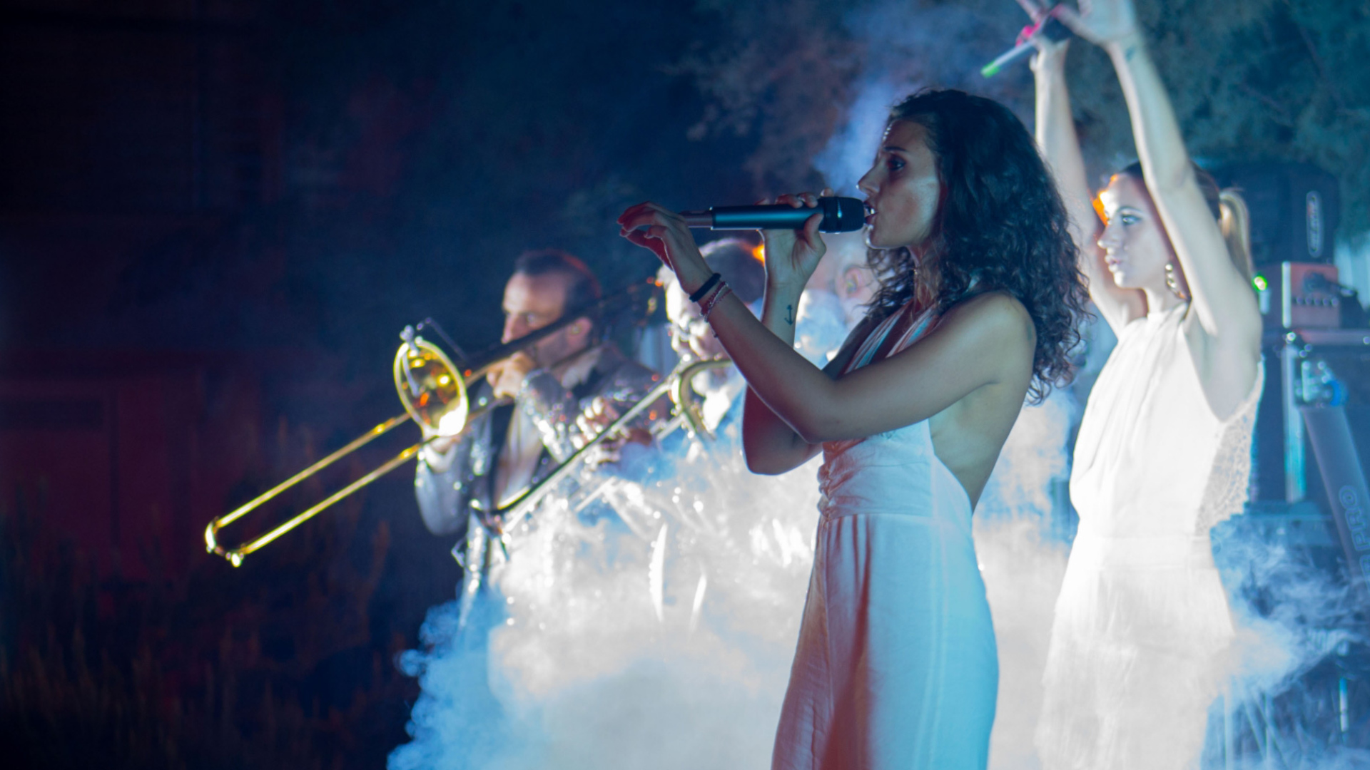 Band musicale dal vivo con cantante e trombonista, atmosfera con fumo e luci.