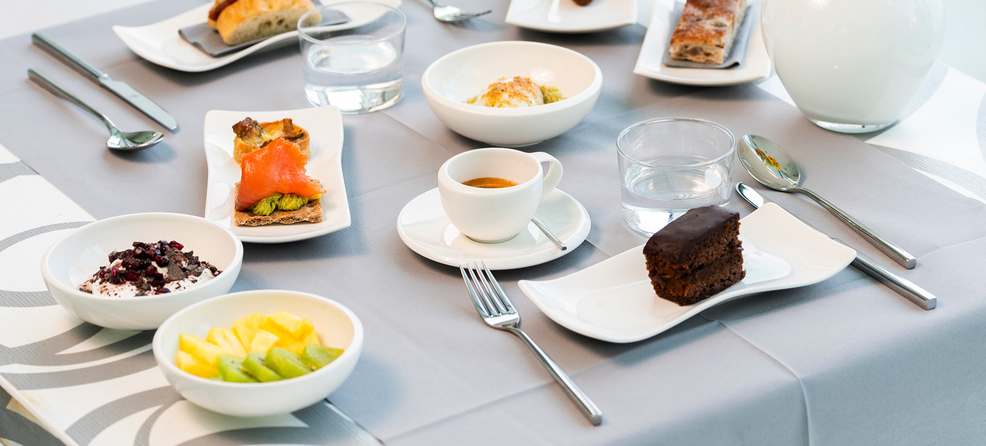 Colazione elegante con dolci, frutta, caffè e acqua su tovaglia grigia.