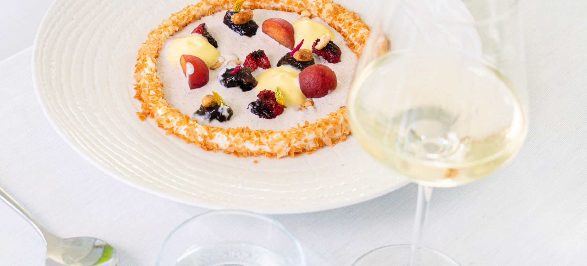 Piatto elegante con frutta, crema e decorazioni, accompagnato da un bicchiere di vino bianco.