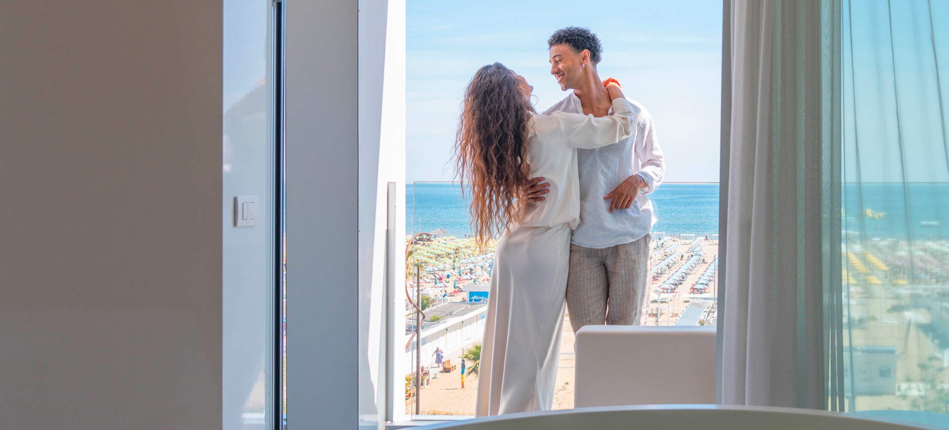 Coppia abbracciata su un balcone con vista mare e spiaggia soleggiata.