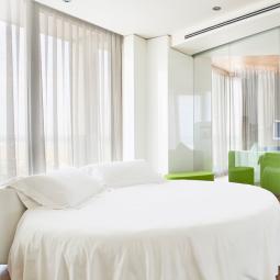 Camera moderna con letto bianco, pareti di vetro e divani verdi.