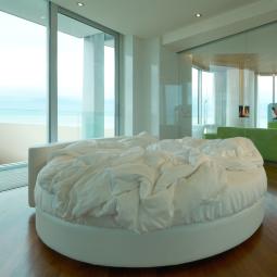 Camera moderna con letto rotondo, pareti di vetro e vista sul mare.