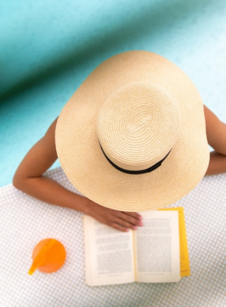 Persona con cappello di paglia legge un libro vicino a una piscina.