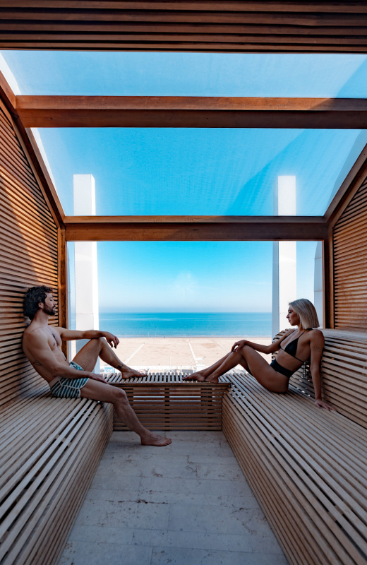 Due persone rilassate in sauna con vista mare, luce calda e atmosfera serena.