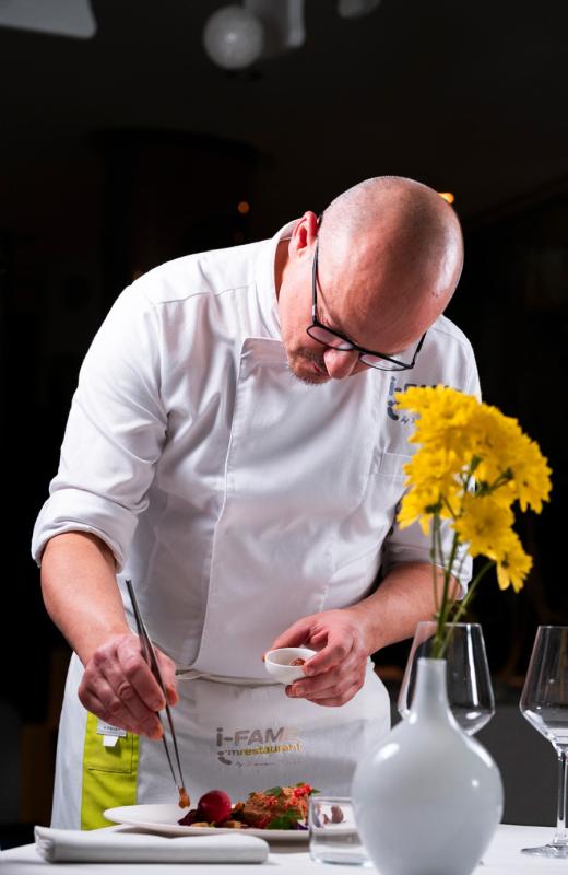 Chef concentrato decora piatto gourmet; fiori gialli sul tavolo, atmosfera elegante e raffinata.