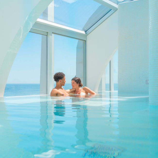 Coppia rilassata in piscina coperta con vista mare e architettura moderna.
