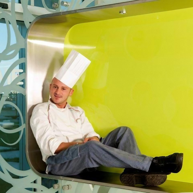 Cuoco seduto in un ambiente moderno con pareti di vetro e design colorato.