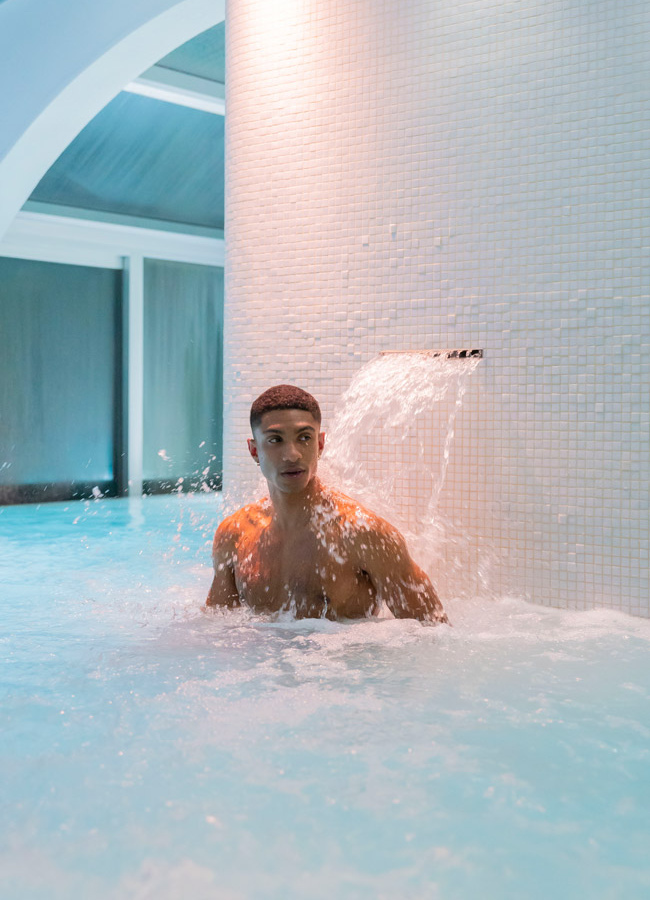 Uomo in piscina, sotto cascata d'acqua, atmosfera rilassante e rinfrescante.