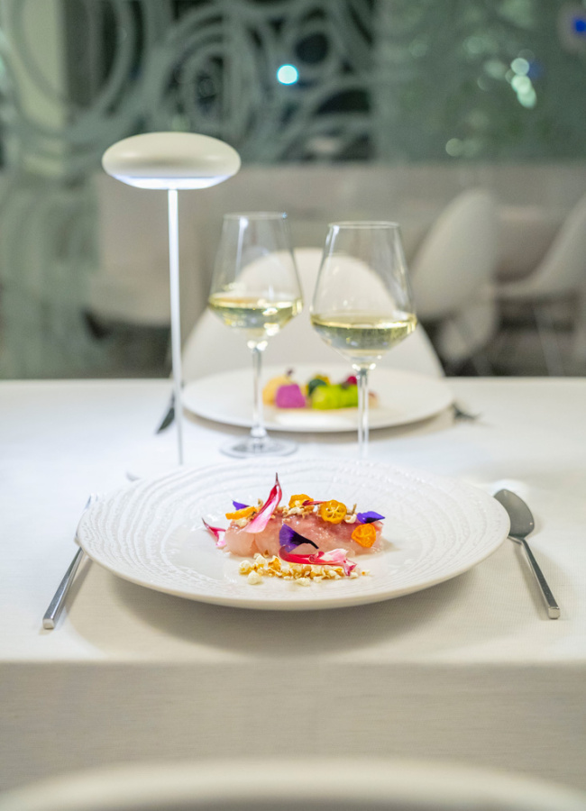 Piatto gourmet con decorazioni floreali, accompagnato da due calici di vino bianco.