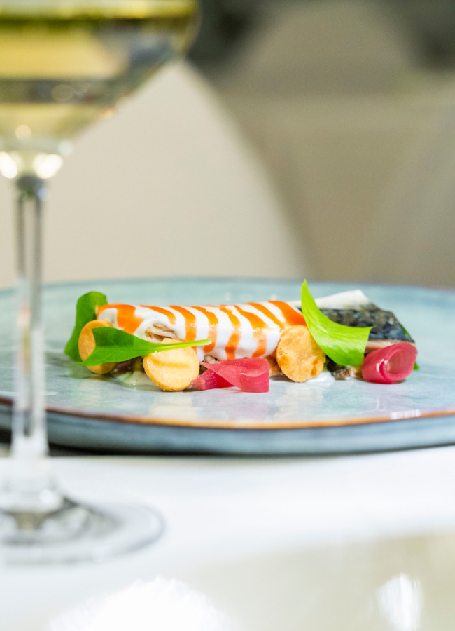 Piatto gourmet con verdure colorate e decorazioni eleganti, accompagnato da un bicchiere di vino.
