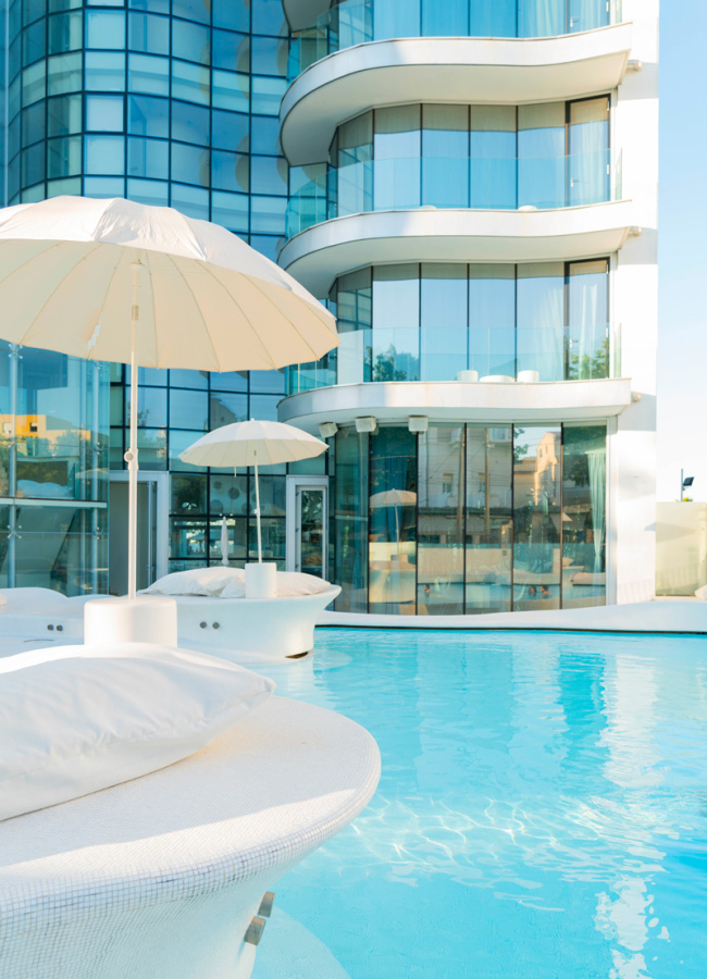 Piscina moderna con lettini e ombrelloni davanti a un edificio di vetro curvo.