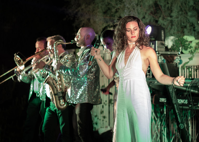 Cantante in abito bianco e band suonano con passione, atmosfera elegante e coinvolgente.