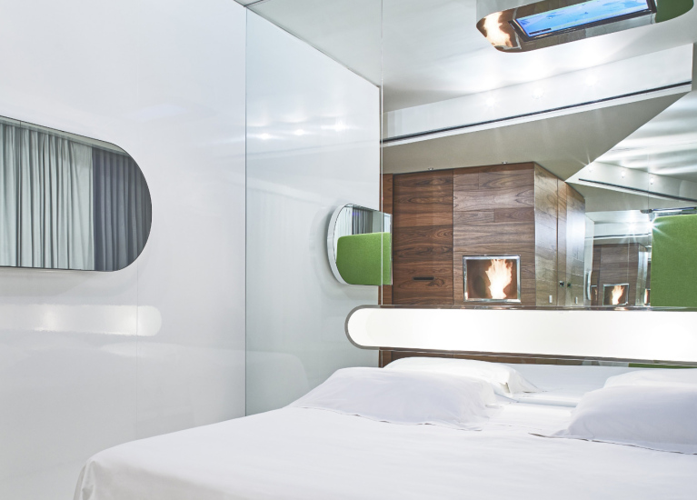 Camera moderna con letto bianco, pareti riflettenti e dettagli in legno e verde.