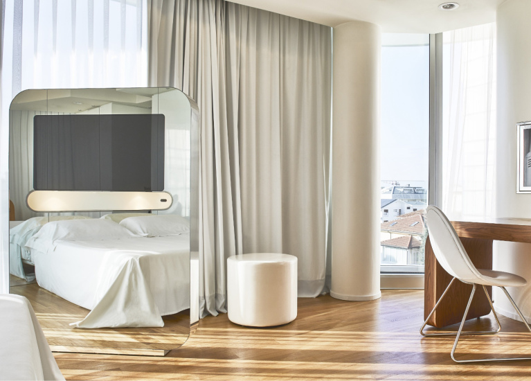 Camera moderna con letto, scrivania di legno e grande specchio, illuminata dalla luce naturale.