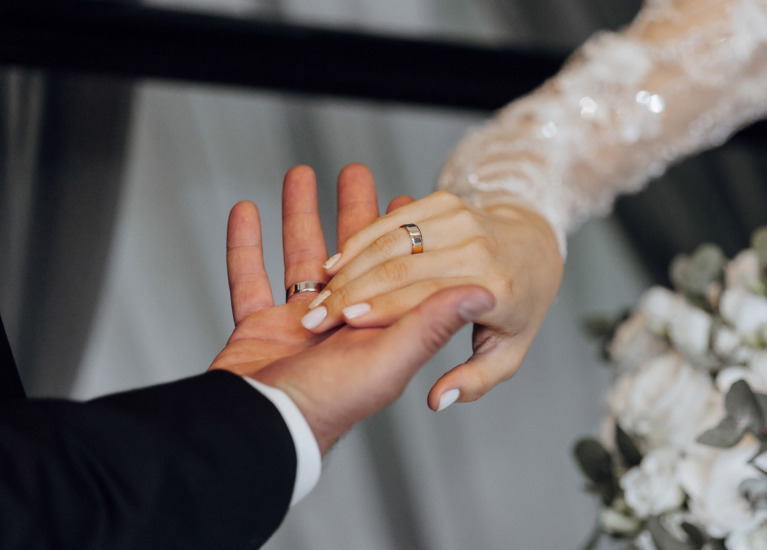 Scambio di anelli tra sposi, mani intrecciate e bouquet di fiori bianchi.