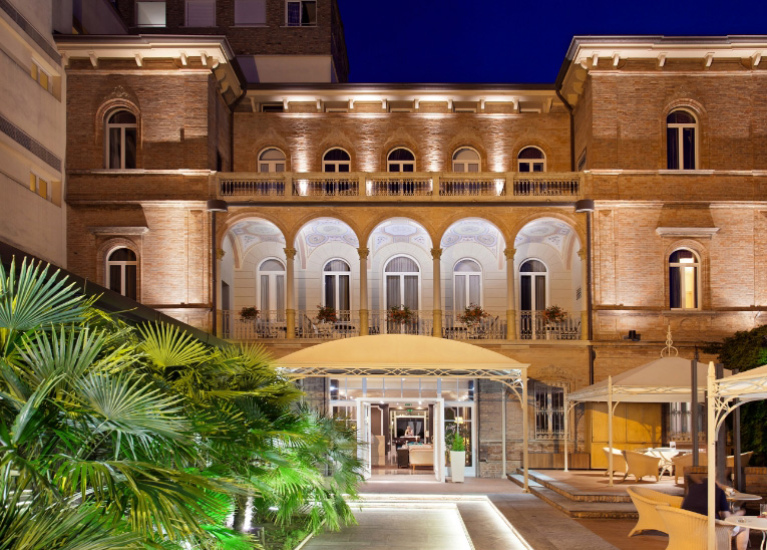 Elegante edificio storico con archi e illuminazione notturna, circondato da vegetazione lussureggiante.