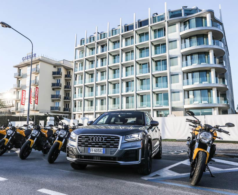Auto Audi e moto Ducati gialle davanti a un moderno hotel, atmosfera di lusso e avventura.