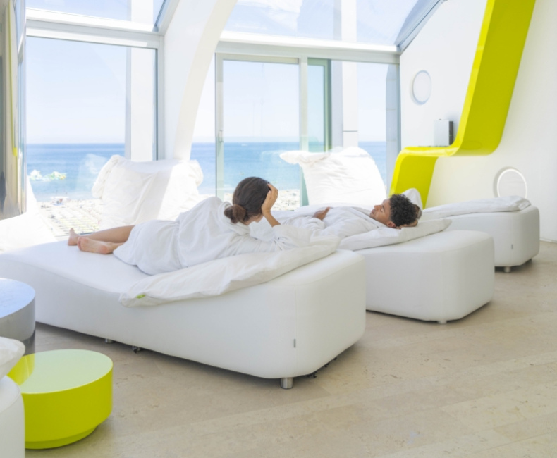 Interno luminoso con vista mare, persone rilassate su letti bianchi moderni.