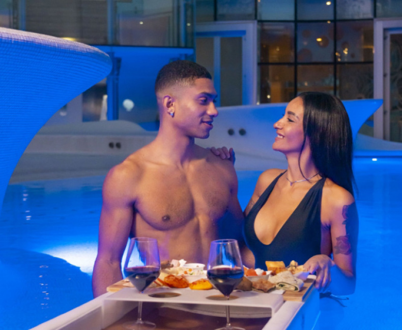 Coppia in piscina con cena galleggiante e bicchieri di vino, atmosfera romantica.