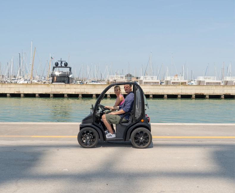 Coppia in mini auto elettrica vicino al porto con barche a vela sullo sfondo.