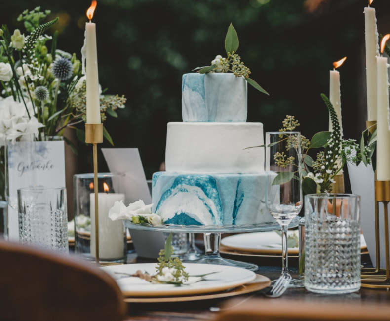 Torta a tre piani blu e bianca su tavola elegante con candele e fiori.